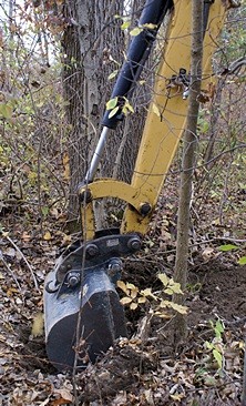 The excavator positioned in the woods digging the large root out from in front of the discharge end of the culvert under the road.