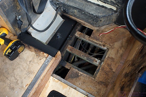 Bus floor under the driver’s seat.  Black tray to rear (upper left) has been sprayed with rubber undercoating.  Seat mounting rails (center) are visible.  Open area is the bay below the driver’s seat.